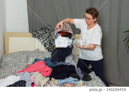 A 52-year-old woman pours clean laundry onto her bed. She is sorting clothes into different piles to organize them. This is happening inside her home in a relaxed setting A 52-year-old woman pours clean laundry onto her bed. She is sorting clothes into different piles to organize them. This is happening inside her home in a relaxed setting 140289459