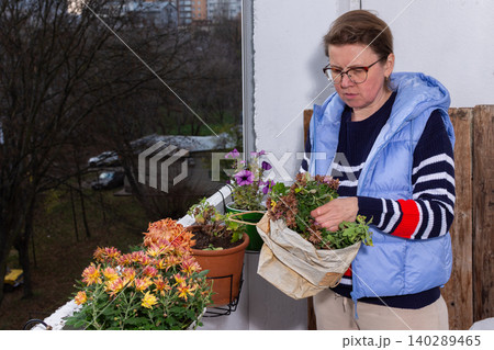 A 50-year-old woman tends to her autumn flowers on a small balcony. She sorts plants from a bag while enjoying her time outdoors in a city environment A 50-year-old woman tends to her autumn flowers on a small balcony. She sorts plants from a bag while enjoying her time outdoors in a city environment 140289465