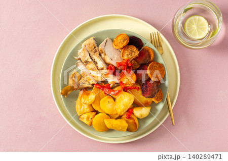 A plate holds baked potato, sliced baked chicken breast, and roasted vegetables like carrots, peppers, and beets. A drink sits nearby with a slice of lime A plate holds baked potato, sliced baked chicken breast, and roasted vegetables like carrots, peppers, and beets. A drink sits nearby with a slice of lime 140289471