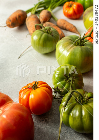 Various types of tomatoes and carrots are placed together on a flat surface. The assortment shows green and ripe tomatoes along with leafy greens and roots 140289472