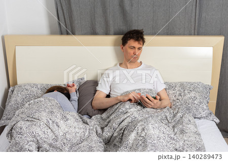 A married wakes up in bed at home. One partner sits up looking at a phone while the other sleeps. They are surrounded by blankets and pillows in a peaceful room A married wakes up in bed at home. One partner sits up looking at a phone while the other sleeps. They are surrounded by blankets and pillows in a peaceful room 140289473
