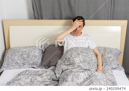 A 53-year-old man wakes up in his bedroom. He is sitting on his bed and holding his forehead with a look of concern. The room has blankets and a gray backdrop 140289474
