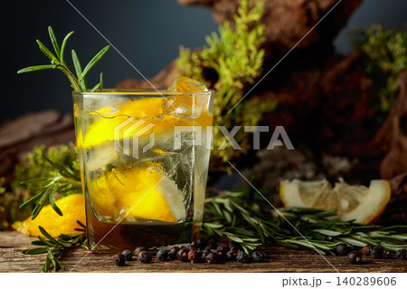 Gin and tonic cocktail with ice, lemon, juniper berries and rosemary sprigs sits on an old weathered board. 140289606