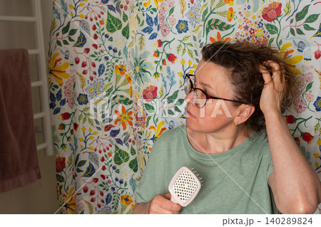 A 50-year-old woman holds a hairbrush while looking in the mirror during her morning bathroom routine. She checks her hair near a floral shower curtain A 50-year-old woman holds a hairbrush while looking in the mirror during her morning bathroom routine. She checks her hair near a floral shower curtain 140289824
