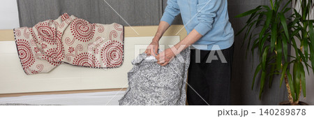A 53-year-old man is changing the bed linen in his bedroom. He holds a pillow while arranging the sheets and blankets, focusing on the task of keeping his space tidy, banner 140289878