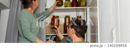 Married places jars of canned goods onto shelves in their pantry. They are inside their home with a view of flowers and the city from the balcony. It is autumn, banner 140289958