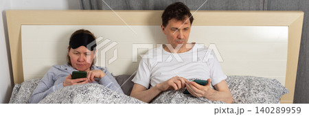 A fifty-year-old wakes up in their bedroom. Both are in bed with blankets. They are looking at their phones in the quiet of the morning, enjoying a relaxed start to the day, banner A fifty-year-old wakes up in their bedroom. Both are in bed with blankets. They are looking at their phones in the quiet of the morning, enjoying a relaxed start to the day, banner 140289959