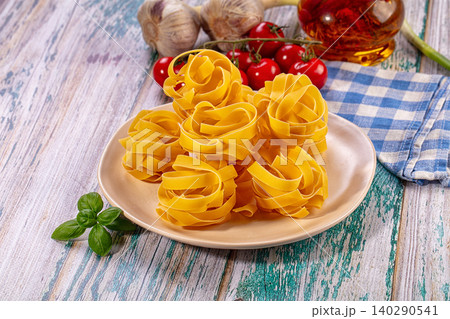 Uncooked tagliatelle pasta nests on beige plate with fresh basil, cherry tomatoes, garlic, and olive oil on rustic wooden surface Uncooked tagliatelle pasta nests on beige plate with fresh basil, cherry tomatoes, garlic, and olive oil on rustic wooden surface 140290541