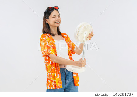 Happy smiling young Asian woman in summer wearing floral shirt holding fan to cool herself with suffers from too hot weather in white background 140291737