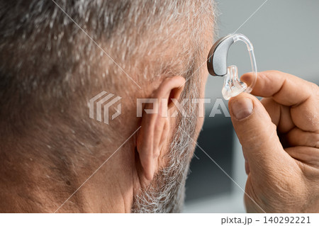 Mature man tries on his custom-made behind-the-ear hearing aid by putting it on his own ear. Hearing solutions for hard of hearing people Mature man tries on his custom-made behind-the-ear hearing aid by putting it on his own ear. Hearing solutions for hard of hearing people 140292221