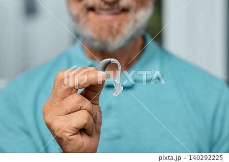 Senior man holding BTE hearing aid in hand on foreground, close-up. Treatment of deafness in elderly people 140292225