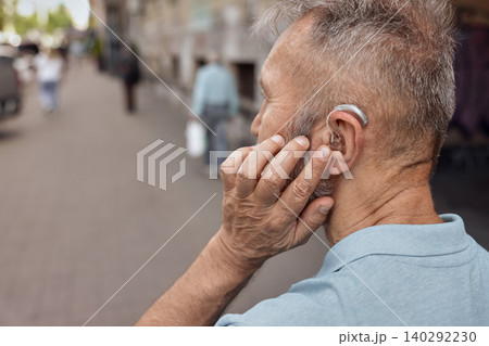 Elderly man with hearing aid behind the ear holds hand near his ear for listening environment urban street, having full life 140292230