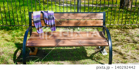 Forgotten sweater draped over a wooden park bench surrounded by green grass and a black fence in a sunny outdoor setting Forgotten sweater draped over a wooden park bench surrounded by green grass and a black fence in a sunny outdoor setting 140292442