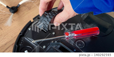 Hand of a technician replacing the wheel motor on a robot vacuum cleaner using a screwdriver, with visible dust and components on a wooden surface 140292593