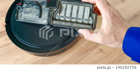 Robot vacuum cleaner maintenance with a hand removing a dirty filter from the device on a wooden floor, showcasing the cleaning process and household environment Robot vacuum cleaner maintenance with a hand removing a dirty filter from the device on a wooden floor, showcasing the cleaning process and household environment 140292756