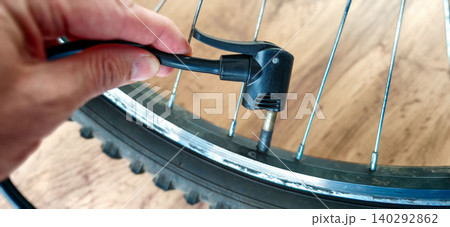 Hand holding bicycle pump nozzle to inflate bicycle tire valve, close-up view of wheel rim and spokes on wooden surface, focus on pump connection Hand holding bicycle pump nozzle to inflate bicycle tire valve, close-up view of wheel rim and spokes on wooden surface, focus on pump connection 140292862