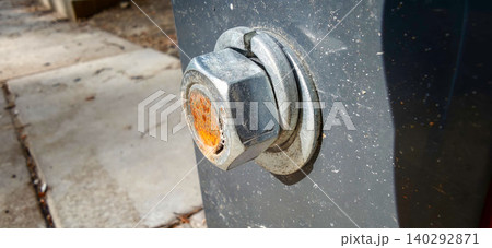 Close-up of a rusty metal bolt and nut attached to a gray surface, showcasing wear and corrosion in an outdoor setting with concrete ground visible 140292871