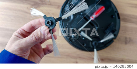 Hand holding replacement brush for robot vacuum cleaner above black base with visible screw holes and screwdriver on wooden surface 140293004