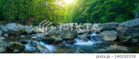 Crystal clear mountain stream flowing among large granite boulders in a lush green forest. Fresh spring landscape with rushing water and sunlit trees in a natural wilderness setting. 140293559