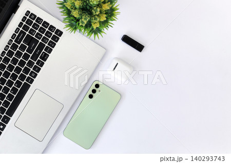 Minimalist workspace with sleek gadgets. Green phone rests beside laptop and mouse. White earbuds and pen add functional touch. Clean desk invites calm, focused productivity 140293743