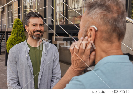 Senior man with a hearing impairment uses a hearing aid to communicate with his male adult friend outdoor. Hearing solutions 140294510