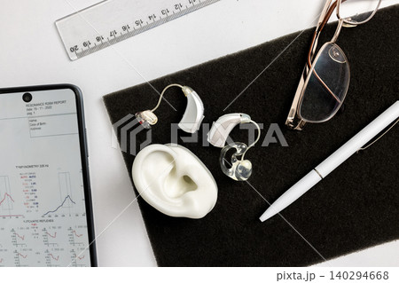 Audiology concept. Advanced hearing aids are featured on a minimalist surface with glasses and a notepad, highlighting modern tech and business style Audiology concept. Advanced hearing aids are featured on a minimalist surface with glasses and a notepad, highlighting modern tech and business style 140294668