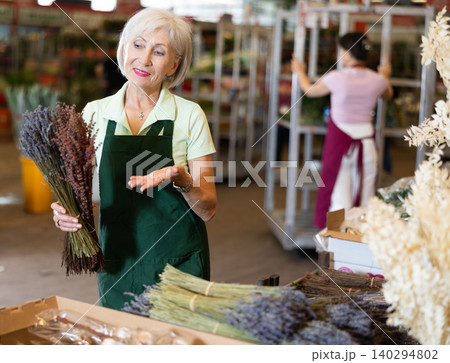 Mature female florist offering bundles of blooming lavender in market Mature female florist offering bundles of blooming lavender in market 140294802