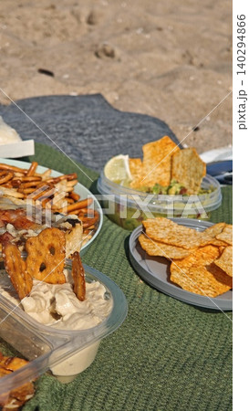 Assorted snacks like pretzels, crackers, and hummus are displayed in containers on a green blanket at the beach, with sand and sunlight visible Assorted snacks like pretzels, crackers, and hummus are displayed in containers on a green blanket at the beach, with sand and sunlight visible 140294866