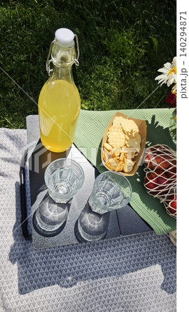 Two glasses of water are placed next to a bottle of yellow drink and a plate of cookies on a picnic table surrounded by green grass and flowers 140294871