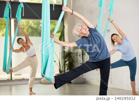 Senior woman practicing aerial yoga asana during group class in gym Senior woman practicing aerial yoga asana during group class in gym 140295012