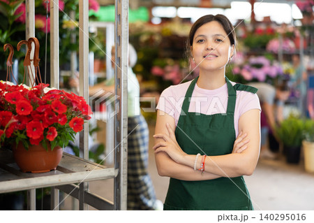 Middle-aged saleswoman standing next to shelves with red petunia in open-air plants market Middle-aged saleswoman standing next to shelves with red petunia in open-air plants market 140295016