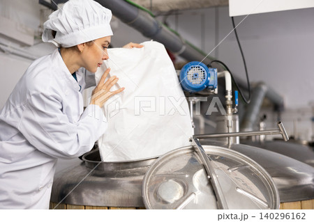Female brewer pouring malted grain from bag into fermenter in brewery 140296162