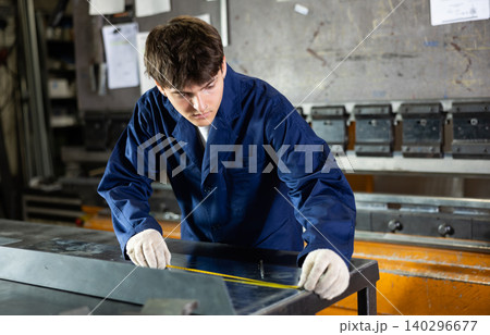 Guy working at metallurgical plant carefully measures sheet of iron using tape measure 140296677