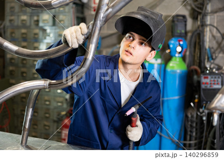 Portrait of male welder standing with welding machine and safety helmet in metal machining workshop Portrait of male welder standing with welding machine and safety helmet in metal machining workshop 140296859