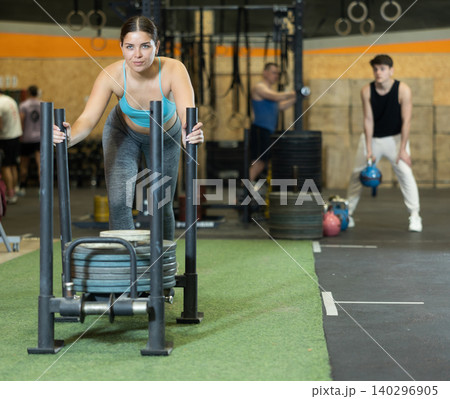 Positive fit young European female in sportswear pushing heavyweight plates on prowler slider in CrossFit center indoors Positive fit young European female in sportswear pushing heavyweight plates on prowler slider in CrossFit center indoors 140296905