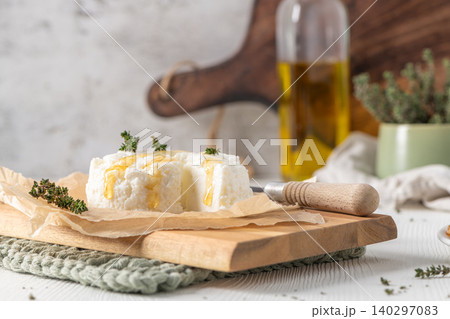 A rustic scene of fresh white ricotta cheese topped with golden honey and thyme sprigs resting on parchment paper and a wooden board with olive oil and kitchen tools in the soft blurred background 140297083