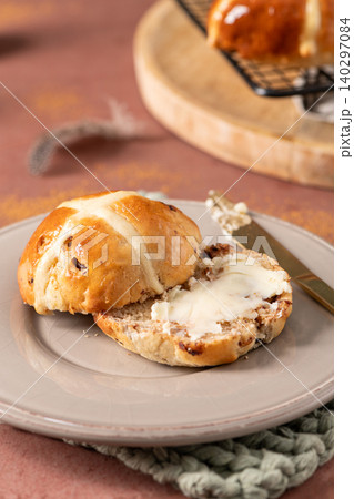 Vertical close-up of a sliced hot cross bun spread with melting butter on a grey plate with a gold knife in a rustic spring kitchen setting Vertical close-up of a sliced hot cross bun spread with melting butter on a grey plate with a gold knife in a rustic spring kitchen setting 140297084