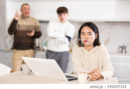 Disgruntled young woman trying to work on laptop while her husband and father scold and shout at her 140297182