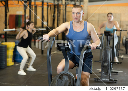 Dedicated active strong middle-aged man doing cardio workout on stationary bicycle as hard cross-training in gym indoors 140297387