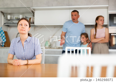 Upset woman sitting in kitchen with angry husband and teenage daughters standing in back Upset woman sitting in kitchen with angry husband and teenage daughters standing in back 140298071