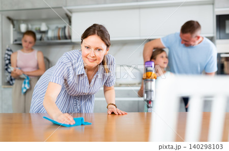 Wife cleans table with rag, husband and children clean kitchen in background Wife cleans table with rag, husband and children clean kitchen in background 140298103