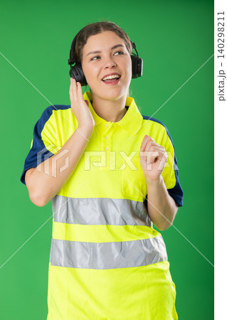 Female worker in reflective polo shirt enjoys listening to music on headphones and dancing on green background Female worker in reflective polo shirt enjoys listening to music on headphones and dancing on green background 140298211