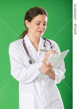 Young woman doctor in white coat with a stethoscope conducts a survey remotely green background 140298241