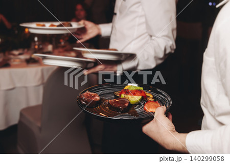Waiter in uniform serving dishes and drinks on party, server carrying plates with food and beverages at the table, catering service on wedding reception, personnel in cafe restaurant, staff on event Waiter in uniform serving dishes and drinks on party, server carrying plates with food and beverages at the table, catering service on wedding reception, personnel in cafe restaurant, staff on event 140299058