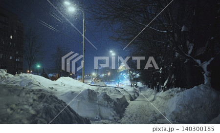 Snowy night street with huge snowdrifts and bare trees under bright city lights Snowy night street with huge snowdrifts and bare trees under bright city lights 140300185