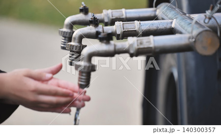 Person washing hands with flowing tap water in a row of outdoor spigots Person washing hands with flowing tap water in a row of outdoor spigots 140300357