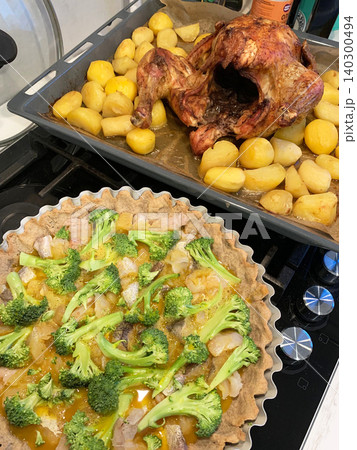 A large baked chicken sits on a tray beside yellow potatoes. A pie filled with white fish and broccoli is in the foreground. This scene shows food being prepared for a festive dinner A large baked chicken sits on a tray beside yellow potatoes. A pie filled with white fish and broccoli is in the foreground. This scene shows food being prepared for a festive dinner 140300494
