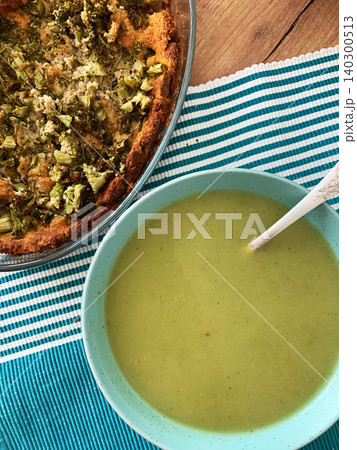 Broccoli cream soup is in a bowl next to a broccoli pie on a table with a striped cloth. The setting is warm and inviting, perfect for a simple meal at home Broccoli cream soup is in a bowl next to a broccoli pie on a table with a striped cloth. The setting is warm and inviting, perfect for a simple meal at home 140300513