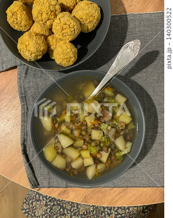 A bowl of soup contains potatoes, broccoli, and green lentils. Next to it, there are baked food balls on a napkin. Both are placed on a wooden table with a gray cloth A bowl of soup contains potatoes, broccoli, and green lentils. Next to it, there are baked food balls on a napkin. Both are placed on a wooden table with a gray cloth 140300522