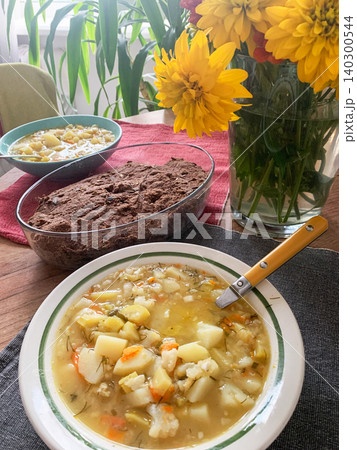 A bowl of soup containing potatoes, zucchini, and cauliflower sits on a wooden table. Fresh bread and yellow flowers are nearby, creating a simple meal scene 140300544